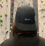 Black five panel hat with black and white American Alpine Club embroidered logo patch worn my model in a climbing gym